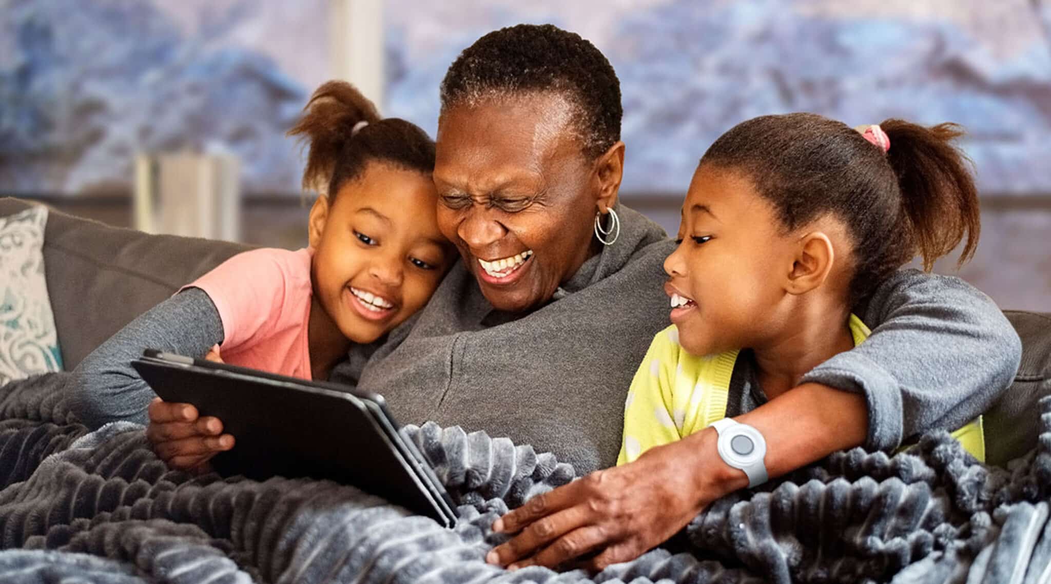 Woman Wearing Medical Alert System Sitting on Couch with Granddaughters Looking at Tablet