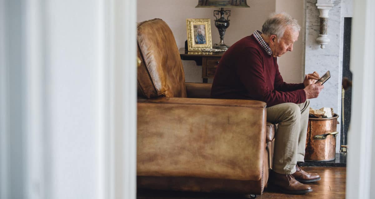 Senior man is sitting in an armchair in the living room of his home, holding and looking at an old photo with a sad expression.